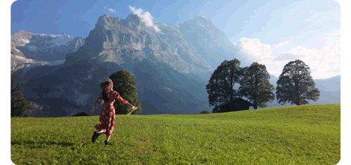 woman in red and white flowery dress jogs through a grass field with trees in the alps