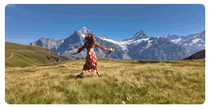 woman in red and white flowery dress jogs through a grass field with trees in the alps
