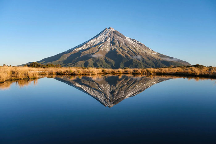 Snow capped mountain covered in brown grass standing in front of clear blue sky and reflection in lake