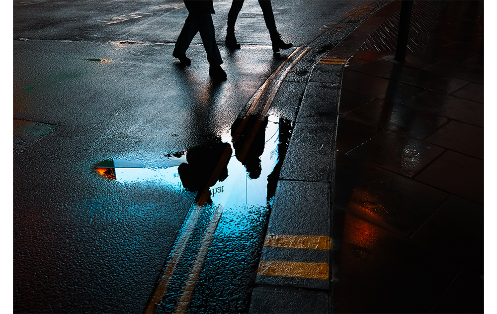fujifilm X100VI sample photograph of London street with puddle reflecting illuminated silhouette of two people crossing the street