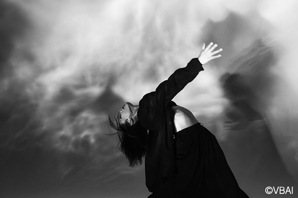 Fujifilm XT5 Acros Film Simulation sample image black and white photo of a woman wearing dark baggy clothing leaning back infront of a whispy smoke cloud