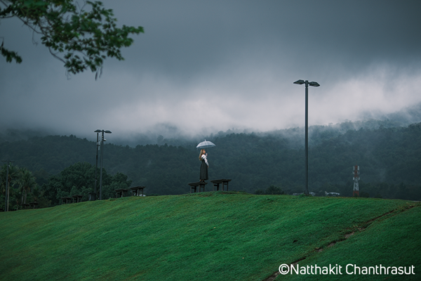 Fujifilm XT5 Classic Negative Film Simulation sample image of a woman standing with an open umbrella at the top of a vibrant green hill with forest and clouds in the background