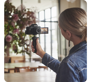female vlogger holding LUMIX G100DV with tripod grip recording selfie video in a light restaurant