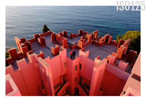 wide angle photograph of pink cubic building with two people standing on roof in front of the ocean