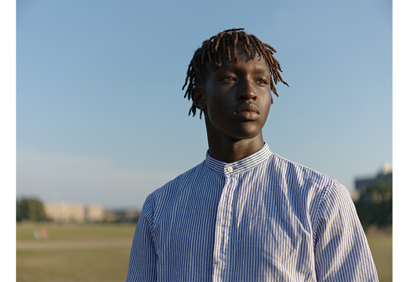 Hasselblad X2D 100C Sample Image of young black man wearing a white and blue pinstriped shirt in an open area with grass and clear blue sky