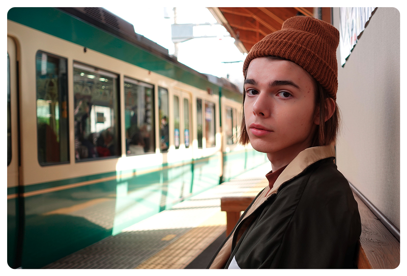 photograph of man wearing brown beanie sat on bench in a trainstation with a green and white train waiting