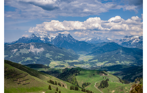 landscape photograph of rolling green hills in the foreground that expand into snow covered mountains in the background with white clouds and bluesky