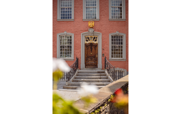 Portrait photograph of a pink brick building with grey stone window surrounds and a open curved entrance staircase leading to a carved wooden door with a golden eagle statuete above