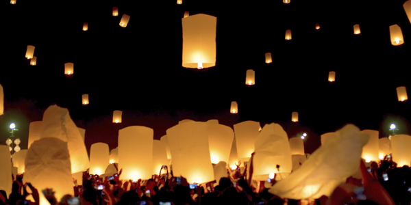 Lit Chinese lanterns floating in a night sky with crowd holding lit lanterns beneath.