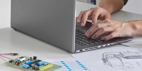 Laptop on a desk with somebody working on it. PCB and paper diagrams scattered on the desk.