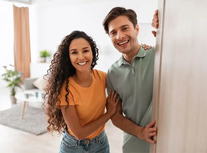 Smiling woman and man, standing closely, beside the doorframe of a living room.