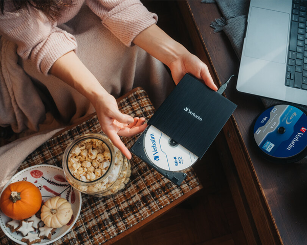 Woman on sofa holding the external blu-ray writer in one hand and a blu-ray disc in the other to highlight its portable convenience