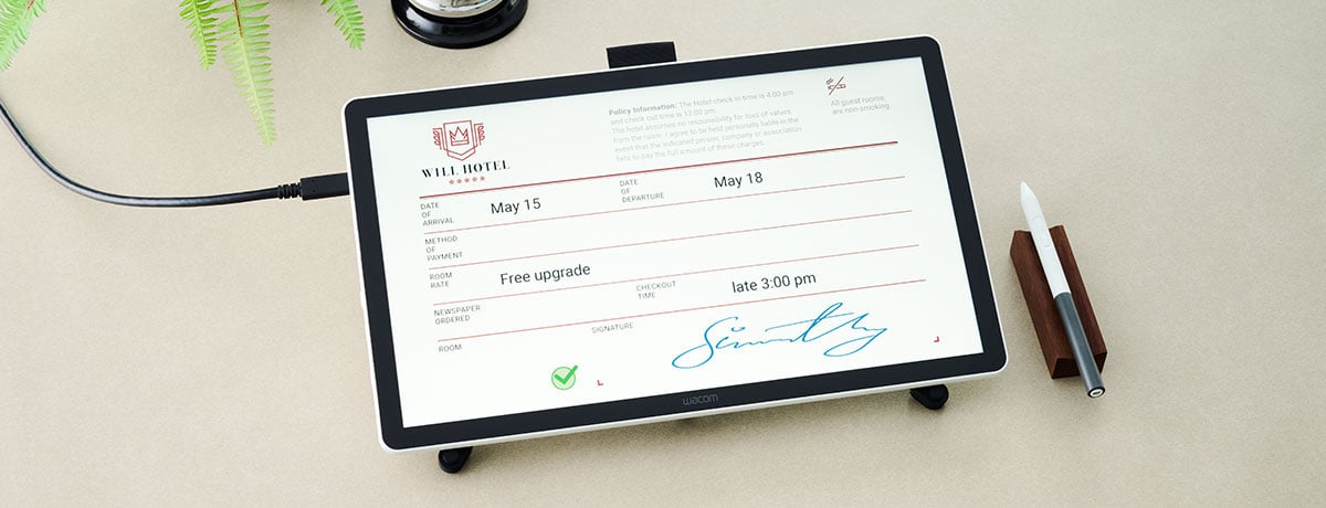 Wacom pen display on a stand showing a digital hotel check-in form with an electronic signature and a stylus on a wooden holder next to it.