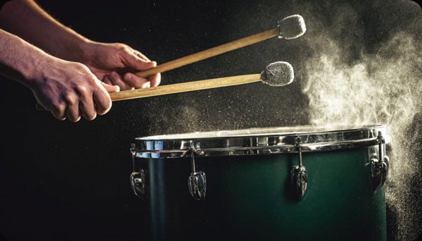 Close-up of a drummer's hands holding mallets above a green snare drum with dramatic smoke and dust effects