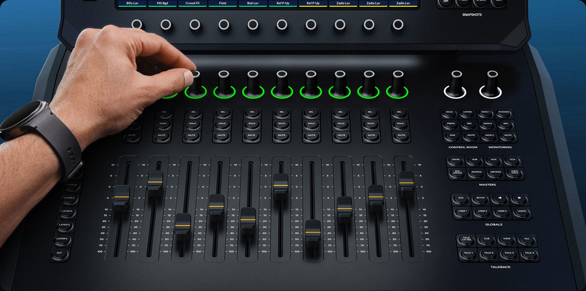 An audio engineer's hand turning a rotary encoder knob on a Blackmagic Design Fairlight Live Audio Panel desktop control surface with motorized faders and glowing green LED ring indicators