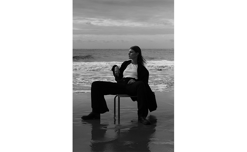 Fujifilm Fujinon XF56mm Lens sample image of female model wearing black suit with white t shirt sat on a chair on a sandy beach with waves behind