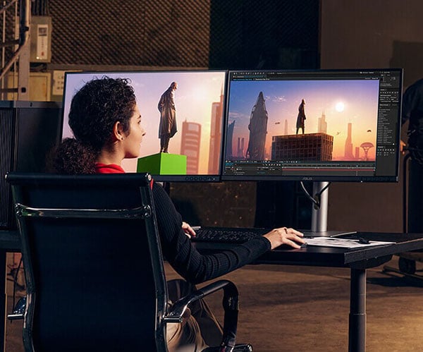 over the shoulder view of a filmmaker sat a desk editing footage on two asus pro art monitors