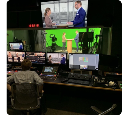 photograph of a chroma key virtual production studio control room with multiple monitors and control panels looking out at two news readers standing infront of a greenscreen