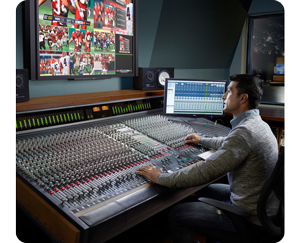 A man sat working at a audio control console in a recording studio with wall monitors