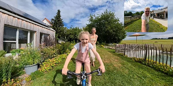 A young girl smiling and riding a bicycle on a grass pathway in a backyard garden with her family