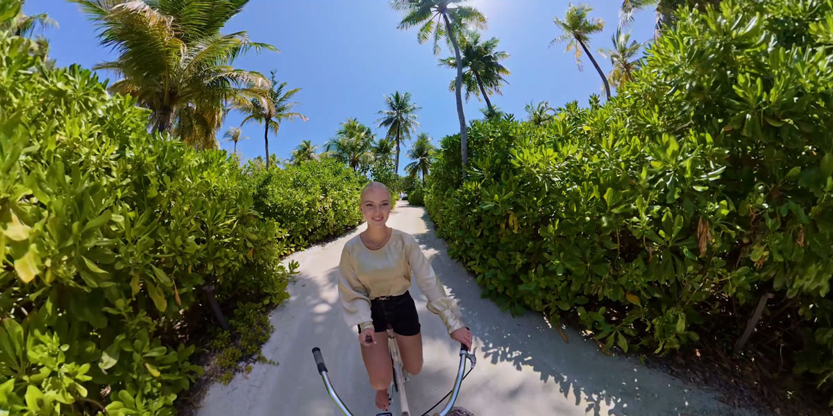A blonde woman riding a bicycle down a sandy path lined with lush tropical greenery and palm trees under a bright blue sky