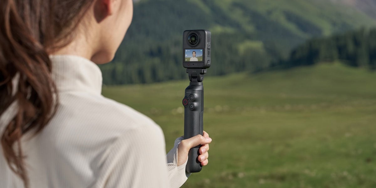 Woman holding a DJI Osmo 360 camera attached to a battery extension rod handle in a green field