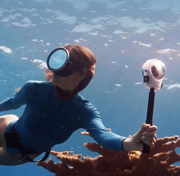 A female scuba diver wearing a vintage oval mask swimming over coral reefs while holding an underwater 360 camera on a selfie stick