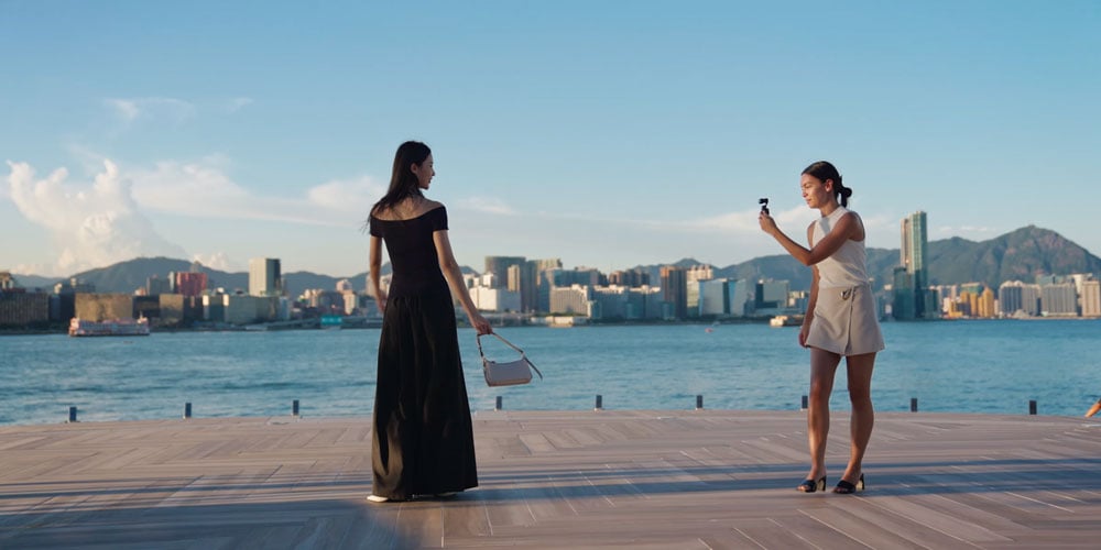 a woman in a black dress being filmed using the dji osmo pocket 4 on a dock with a city in the background
