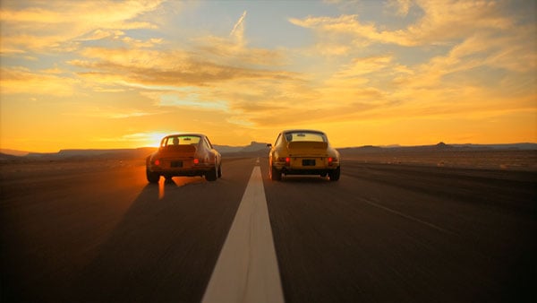 two vintage cars driving down a highway at sunset