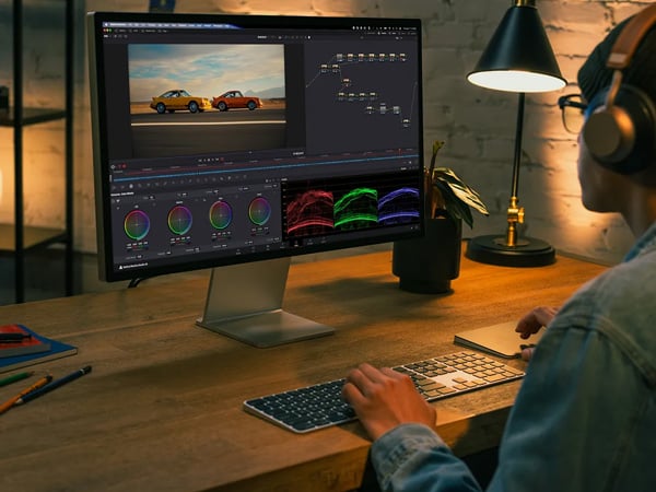 an Apple Studio Display monitor on a wooden desk with a person editing video footage in DaVinci Resolve