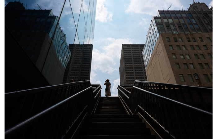 hasselblad X2D XCD 4/28P Sample image of a woman wearing a long dress and hat at the top of double stair set with buildings on both sides with a bright blue and cloudy sky
