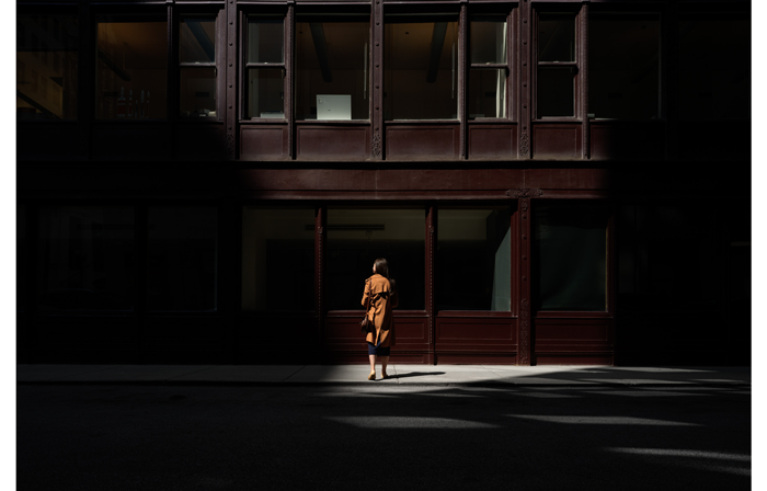hasselblad X2D XCD 4/28P Sample image of a woman wearing a brown rain coat and handbag stood facing away from camera looking a deep maroon coloured building with large windows, draped in shadow and bright sunlight highlighting the woman in frame