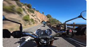 image of motorcycle handle bars with road outstretched in front with another motorcyclist in front with arms outstretched on a coastal tunnel road