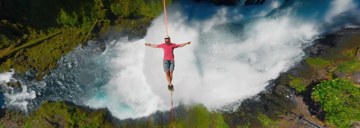 top down view of a man balancing on a highline strung across a roaring waterfall in a forested area