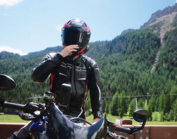 a motorcyclist wearing a dark leather riding jacket and a full-face helmet with red details while seated on a motorcycle against a mountain backdrop with the insta360 x6 maounted to their bike