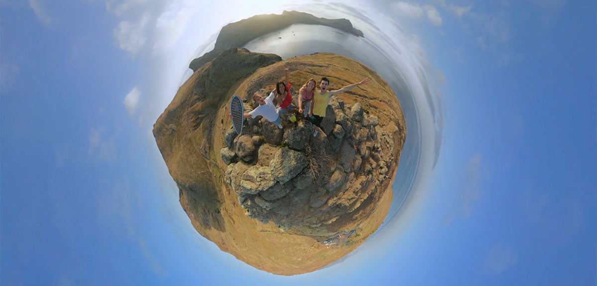 Four people appear to wave on top of a rocky peak with a coastal or island backdrop using a tiny panet shot