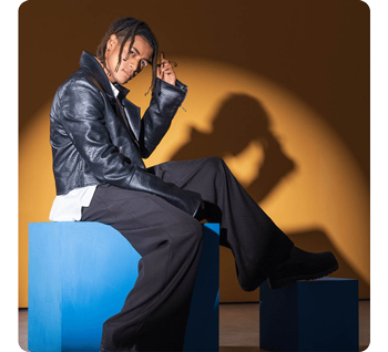 Male model wearing black leather jacket sitting on blue box infront of orange studio wall lit by hard white light