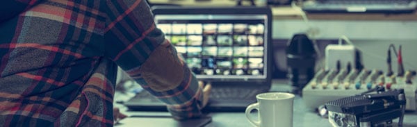 a person in a plaid shirt working at a desk with a laptop displaying a grid of video clips
