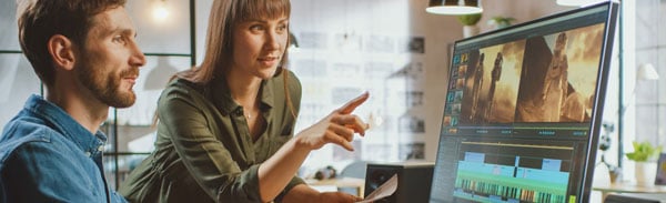 A woman pointing at a computer monitor while discussing video editing software with a male colleague in a modern office or studio