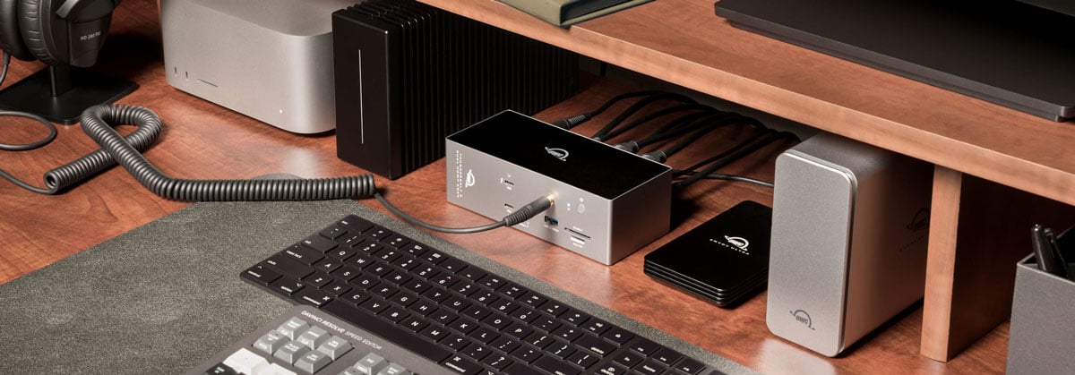 OWC Thunderbolt 5 Dock on a wooden desk setup with a mechanical keyboard, external SSD, and studio monitors under a monitor riser.