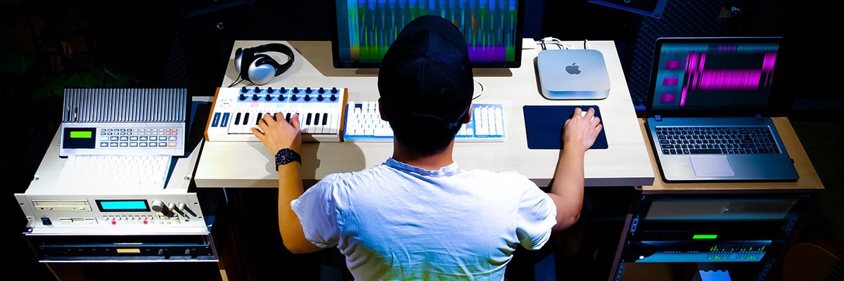 Overhead view of a music producer in a white t-shirt using a MIDI controller, Mac Mini, and laptop in a recording studio