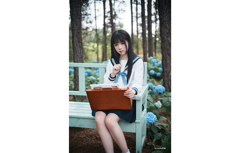 TTArtisan 50mm F2 sample photograph of asian model holding briefcase and pen whilst sitting on wooden park bench in front of blue flowers and trees