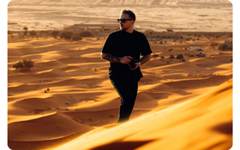 photograph of main wearing black t-shirt and jeans holding camera standing in golden desert sand dunes that expand out behind