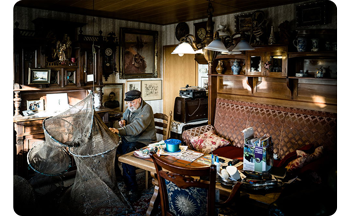 Photograph of old man sat knitting fishing nets in his home surrounded by old wooden furniture