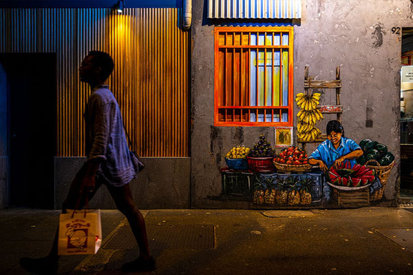 Photograph of woman walking past street art painting of female market vendor selling fruit below orange barred windows
