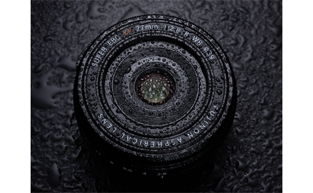 Fujfilm 27mm weather resistant Lens covered in water droplets on a black surface
