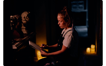 Sample image of femal artist sitting sketching on pad in dark candle lit room surrounded by marble busts