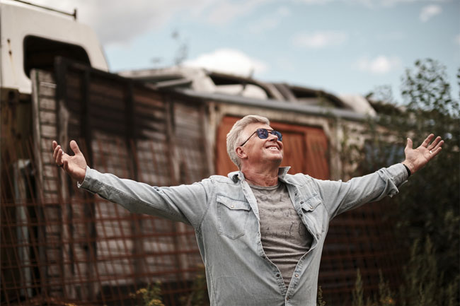 knut koivisto sample image of man wearing denim jacket standing arms outstretched to the sky in front of derelict wooden fence