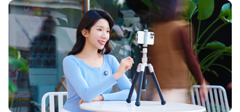 photograph of asian woman wearing blue shirt sitting at table with smartphone and tripod