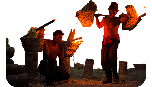 Men carrying mined stone in woven baskets on their shoulders lit by fire light